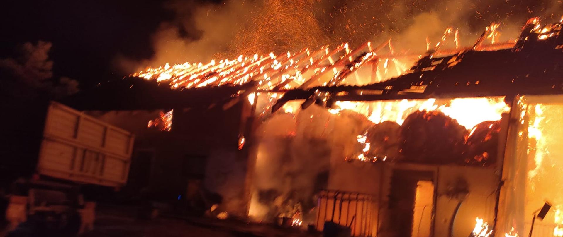 Zdjęcie przedstawia pożar budynku gospodarczego. Jest pora nocna. Na zdjęciu widać że budynek gospodarczy jest cały objęty jest ogniem widać płomienie i dym. Przed budynkiem widać stojącą część przyczepy. 