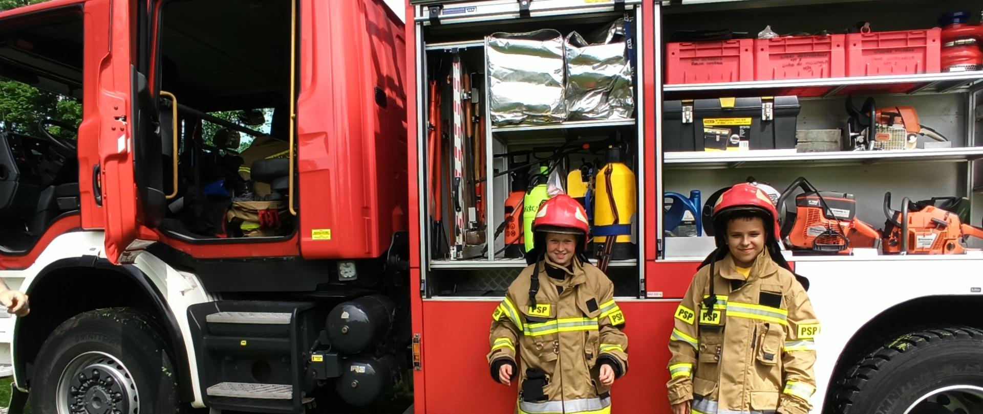 Uczestnicy IV Pikniku Militarnego w kurtkach i hełmach strażackich na tle samochodu ratowniczo-gaśniczego