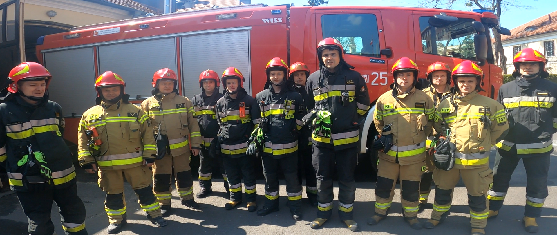 Jednostka ratowniczo-gaśnicza numer 4 , w tle samochód gaśniczy a przed pojazdem stoją strazacy w umundurowaniu bojowym z chełmami na głowach.