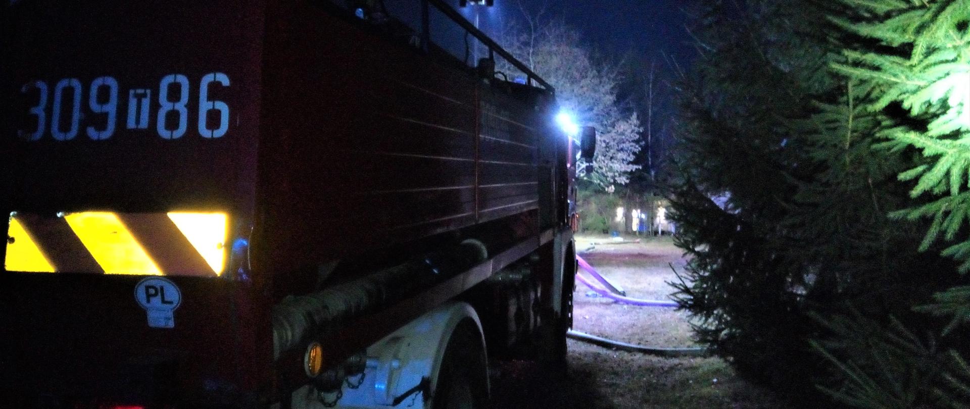 Fotografia przedstawia nocna perspektywę miejsca pożaru. Po lewej stronie zaparkowany samochód gaśniczy, przed nim rozwinięte linie gaśnicze.