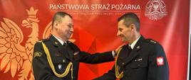 Zdjęcie przedstawia Komendanta Powiatowego PSP przyjmującego życzenia z okazji przejścia na zaopatrzenie emerytalne. W tle czerwony baner Komendy Powiatowej PSP w Wolsztynie.