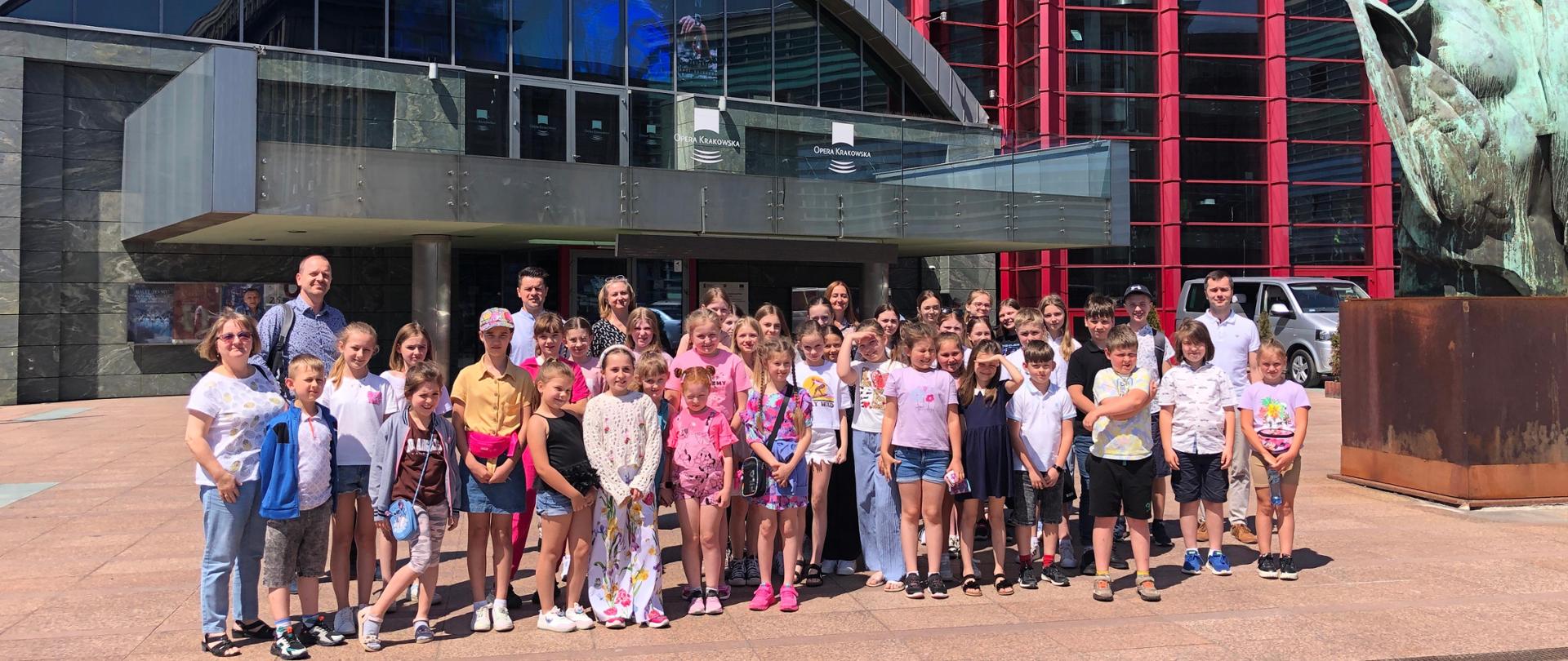 na zdjęciu widnieją uczniowie i nauczyciele PSM I st. w Jaśle, którzy stoją w kilku rzędach przed budynkiem Opery Krakowskiej. Kolorystyka zdjęcia jest szaro-brązowo-czerwono-niebiesko-czarna.