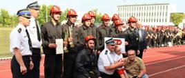 dziewięciu zawodników oraz przedstawiciele władz lokalnych wraz z komendantem powiatowym pozują na stadionie sportowym do pamiątkowej fotografii, w tle murawa boiska oraz pozostałe drużyny zawodników