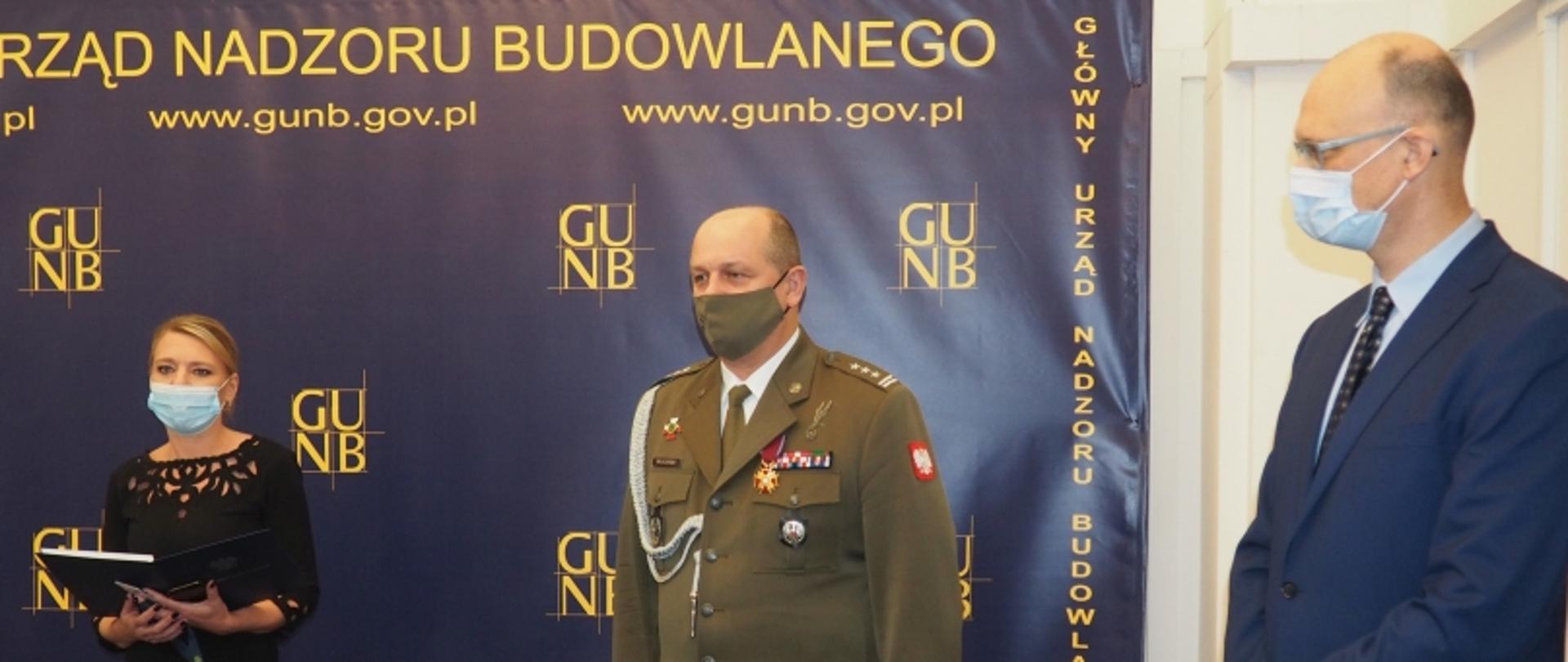 „Trzy osoby przed ścianką z napisem Główny Urząd Nadzoru Budowlanego i adresem www.gunb.gov.pl; jedna w mundurze, jedna z teczką, jedna w garniturze i maseczce.”