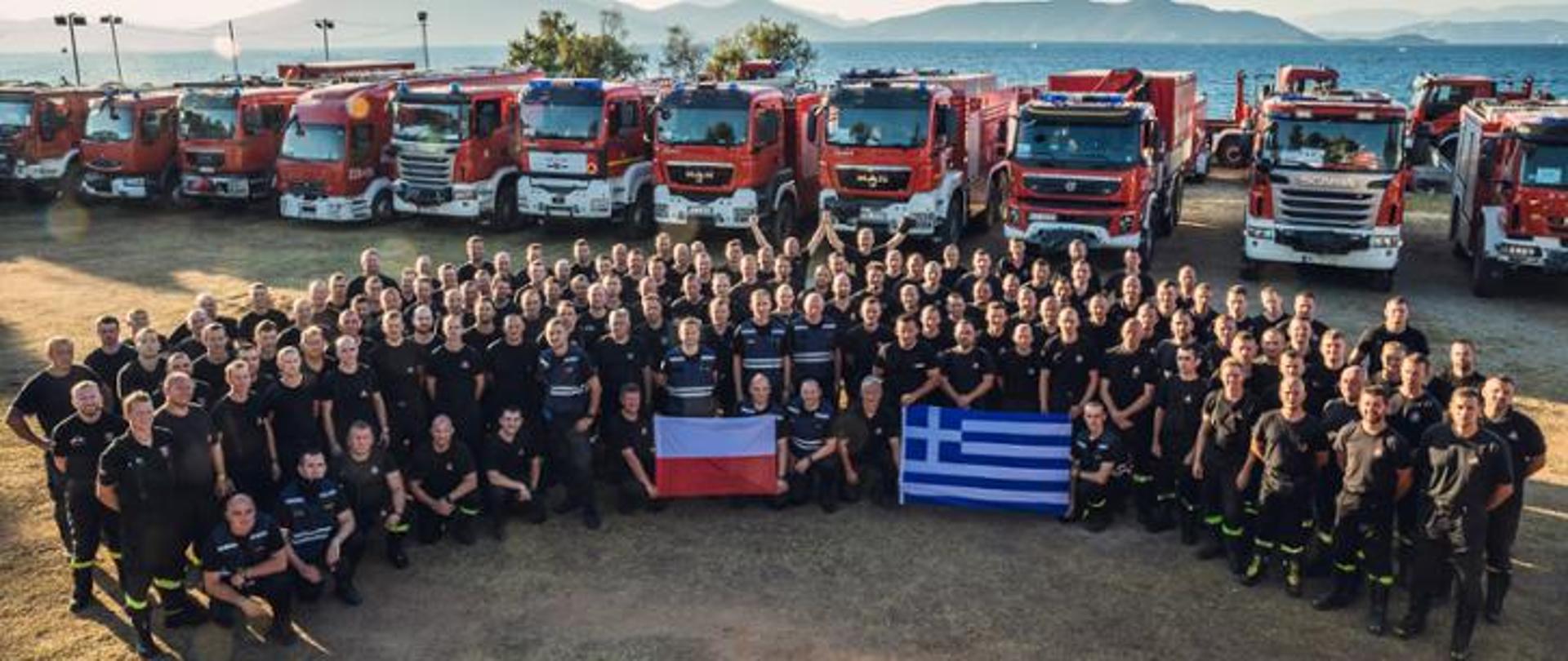 Zdjęcie grupy polskich strażaków na terytorium Grecji. Grupa 143 strażaków z polski oraz kilku strażaków z Grecji trzymają flagi Polski i Grecji. W tle samochody pożarnicze ustawione w kolumnie