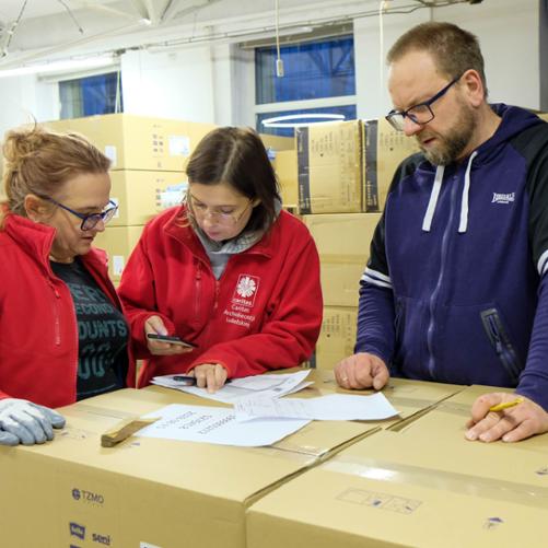 W magazynie pełnym paczek z pomocą materialną obok jednego stosu pudeł stoją dwie kobiety i mężczyzna i patrzą się na leżące na pudłach kartki