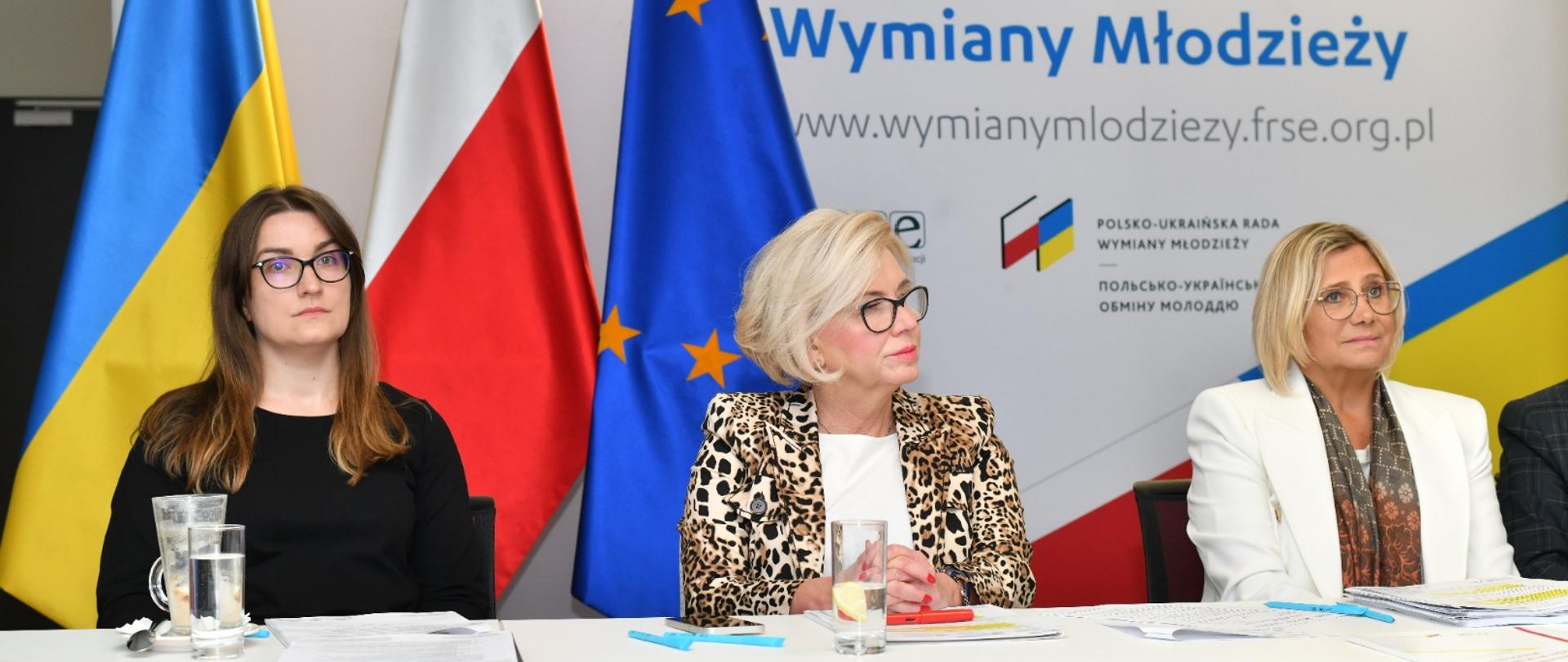Przy białym stole siedzi wiceminister Machałek w marynarce w brązowo-żółto-biały wzorek, kobieta w białej marynarce i kobieta w czarnym ubraniu, za nimi flagi Polski, Ukrainy i UE, za nimi na ściance napis Polsko-Ukraińska Rada Wymiany Młodzieży.