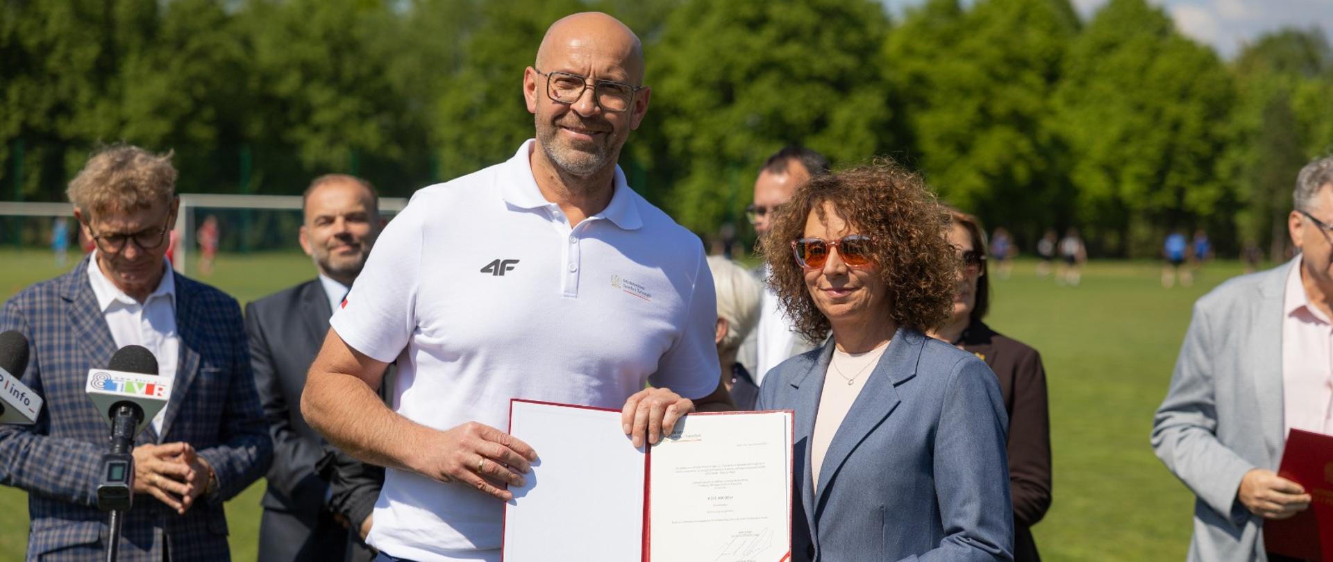 Konferencja prasowa na boisku piłkarskim. Minister sportu i turystyki Jakub Rutnicki i burmistrz Łasku Monika Mrowińska prezentują decyzję MSiT o przyznaniu dofinansowania do budowy pełnowymiarowego boiska piłkarskiego. Za nimi inni uczestnicy konferencji.