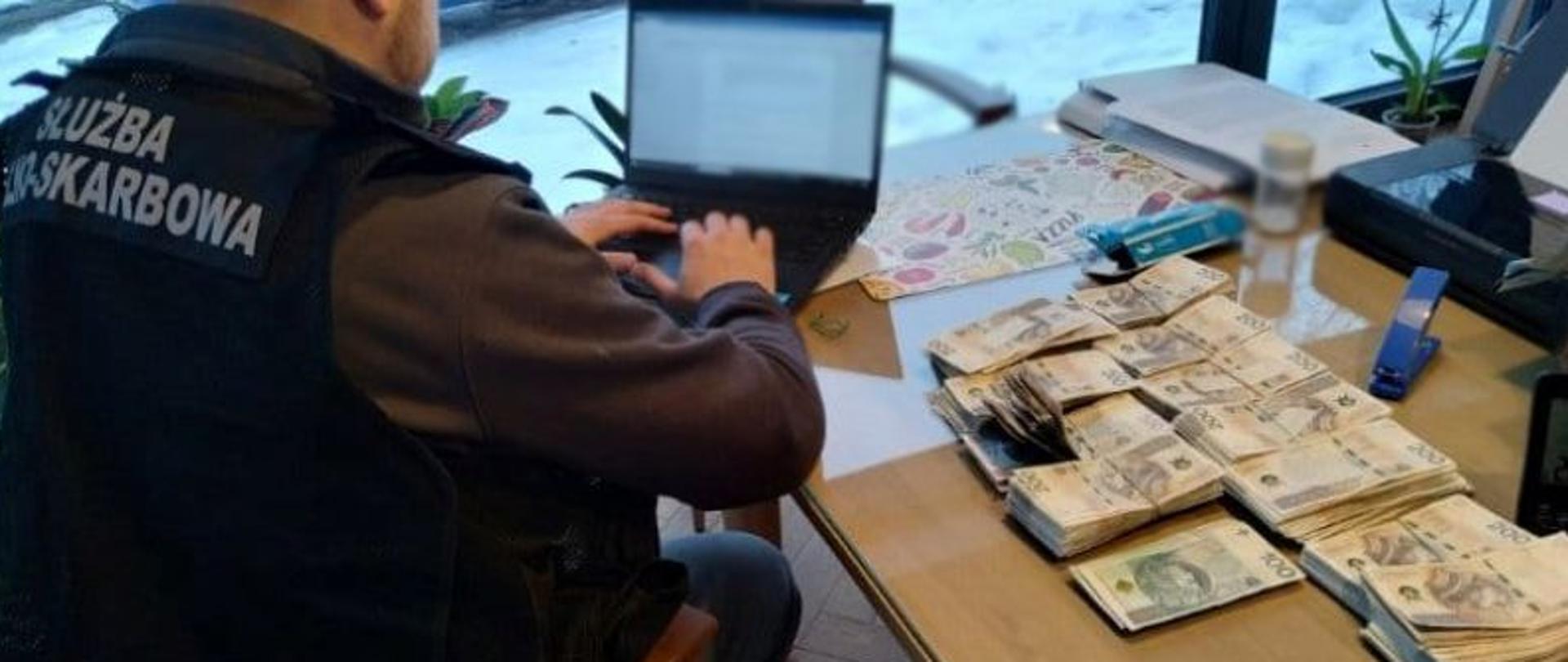 Funkcjonariusz Służby Celno‑Skarbowej pracuje przy komputerze przy stole, na którym leżą duże pliki banknotów.