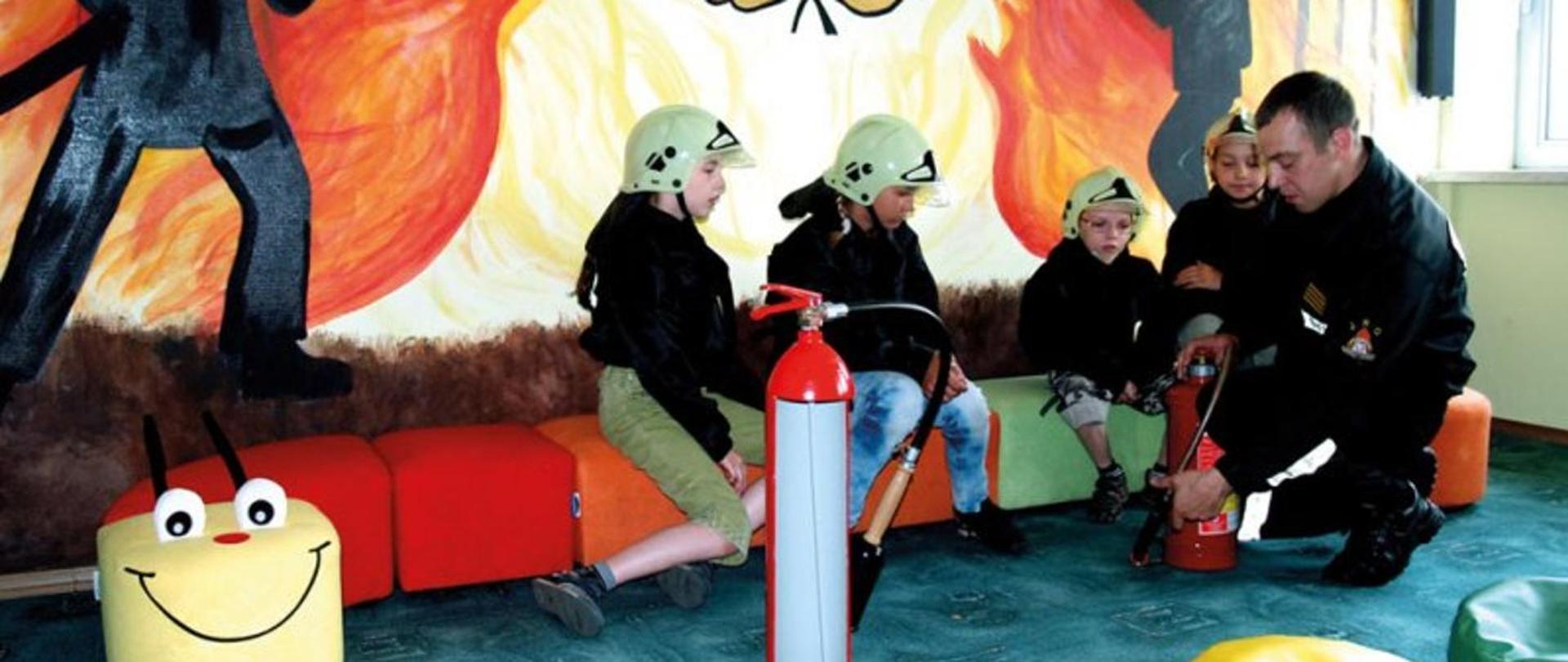 Zdjęcie przedstawia czwórkę dzieci w chełmach, którym strażak pokazuje gaśnicę. 