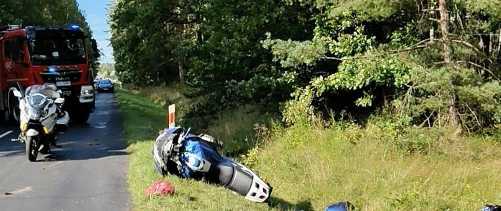 Na poboczu drogi leży przewrócony niebieski motocykl, a wokół niego porozrzucane są elementy kasku i części maszyny. W tle widać wóz strażacki oraz motocykl policyjny. Z lewej strony kadru biegnie asfaltowa droga, a prawa strona to pas zieleni i las. Motocykl jest wyraźnie uszkodzony, leży na boku. Scena przedstawia skutki wypadku drogowego motocyklisty.