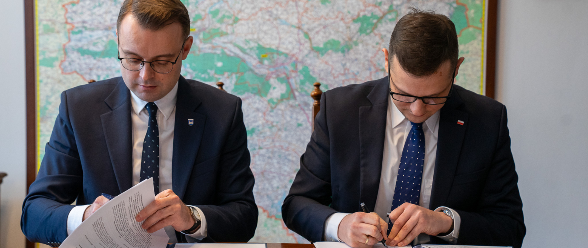 Podpisanie umowy na nowy żłobek w Żninie przez Wojewodę Michała Sztybla oraz Burmistrza Żnina Łukasza Kwiatkowskiego. Za plecami mężczyzn widoczna jest mapa województwa kujawsko-pomorskiego 