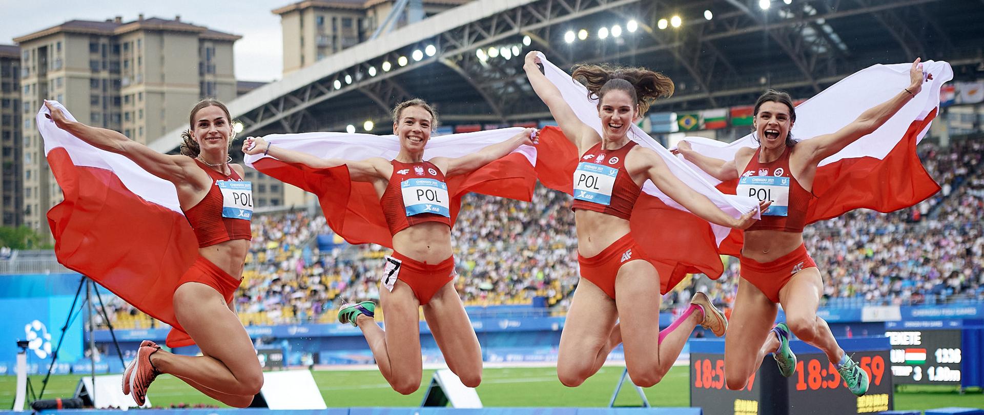 Cztery trzymające biało-czerwone flagi dziewczyny w czerwonych sportowych strojach skaczą nad boiskiem, w tle trybuny.