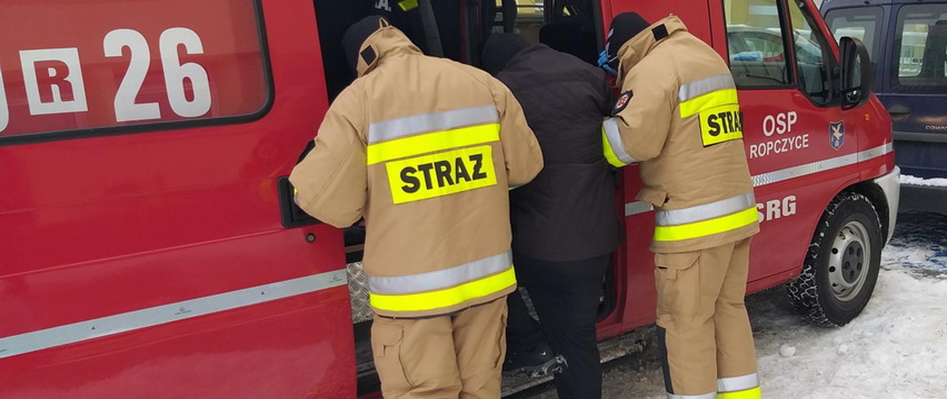 Dwaj strażacy w umundurowaniu bojowym pomagają starszemu mężczyźnie w czarnym płaszczu wsiąść do czerwonego samochodu pożarniczego, trzymając go pod ręce. Mężczyzna jedną nogą stoi na progu samochód typu bus z napisem na przednich drzwiach OSP Ropczyce. Samochód stoi na zaśnieżonym parkingu.
