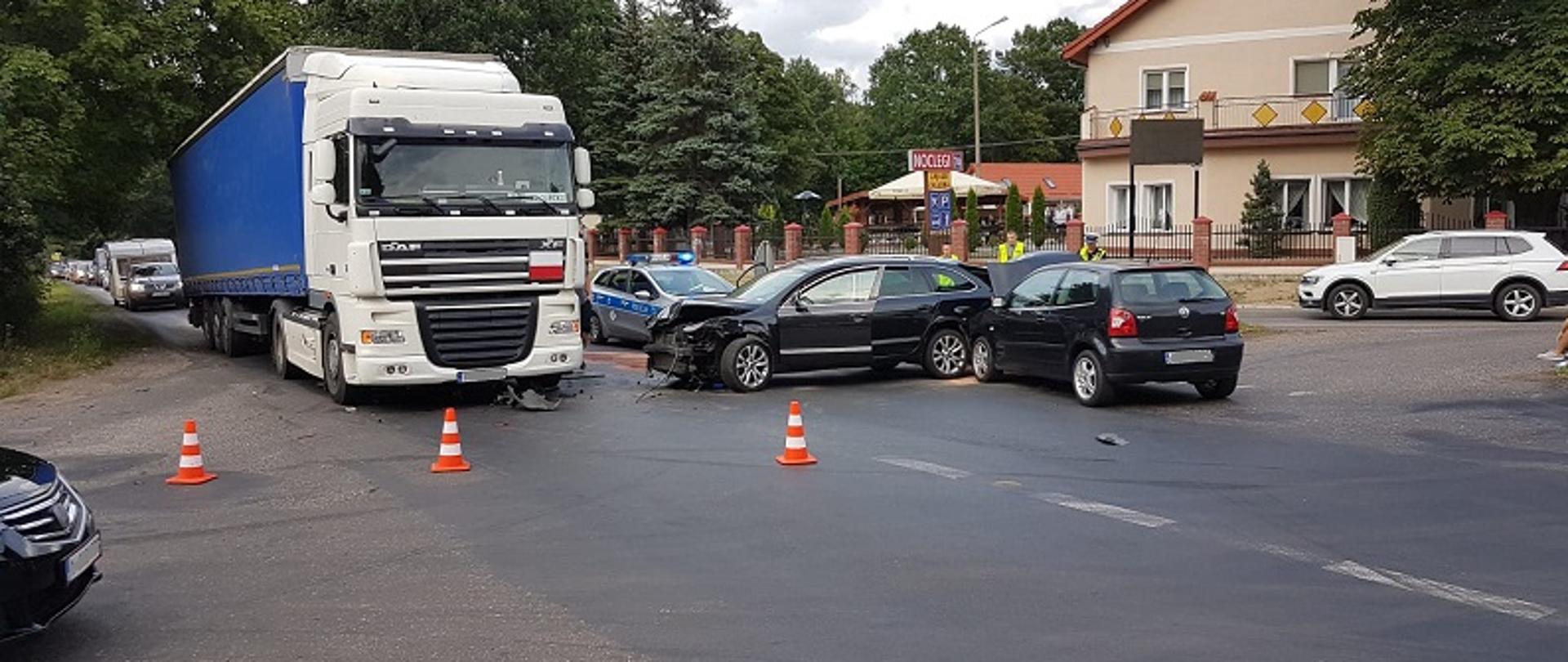 Wypadek w miejscowości Byszyno w powiecie białogardzkim. Zdjęcie przedstawia uczestniczące w zdarzeniu pojazdy na skrzyżowaniu dróg. Przodem widoczny samochód ciężarowy DAF, bokiem od strony kierowcy widoczna Skoda z uszkodzonym pasem przednim, Od tyłu widoczne czarny pojazd marki VW z otwarta maską przednią dotykający tylnej części Skody. Za samochodami policjanci i osoby uczestniczące w wypadku