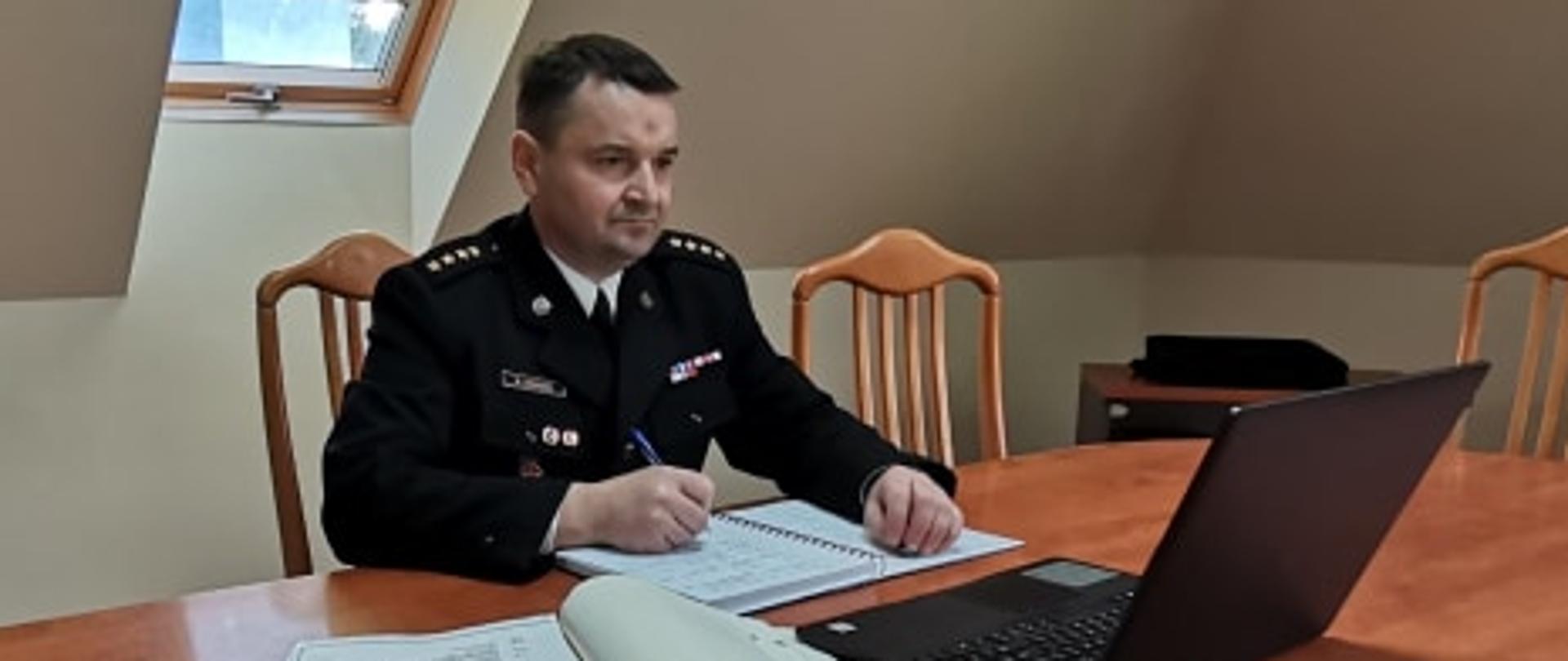 NA ZDJĘCIU WIDAC STRAŻAKA W UMUNDUROWANIU GALOWYM SIEDZĄCEGO PRZY STOLE. NA STOLE LEŻY KOMPUTER PRZENOŚNY I DWA NOTESY , STRAŻAK PATRZY W EKRAN , TRZYMA W PRAWEJ DŁONI DŁUGOPIS.