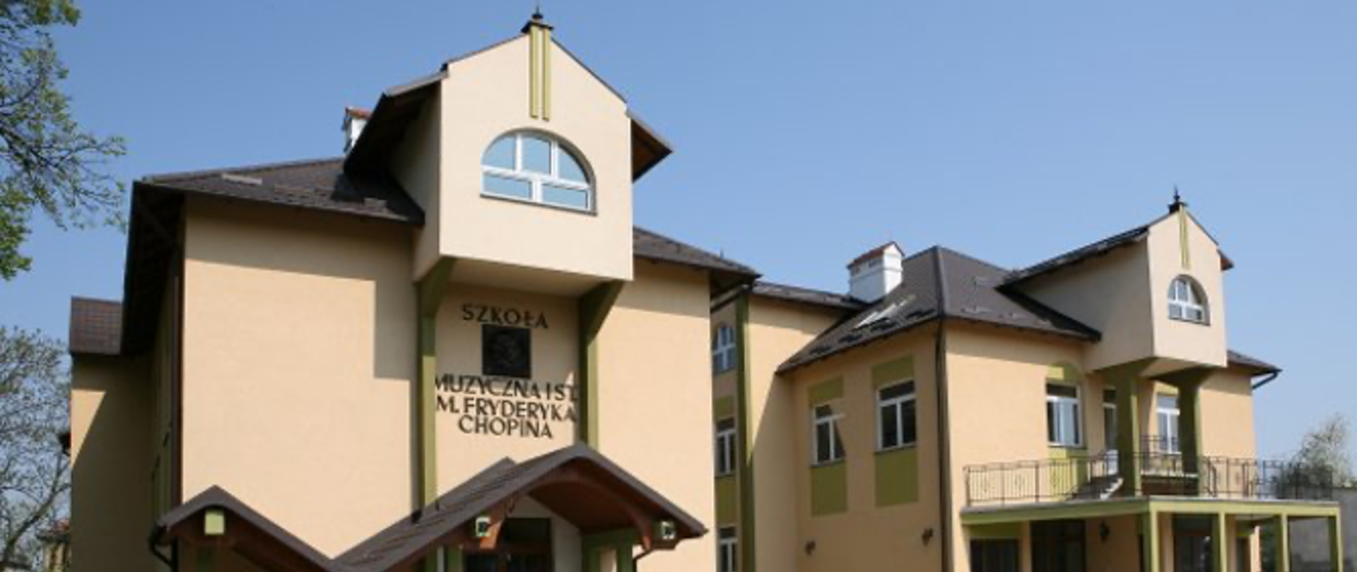 Budynek Szkoły Muzycznej im. Fryderyka Chopina w Jarosławiu, widoczny od frontu w słoneczny, bezchmurny dzień. Jest to dwupiętrowy gmach o jasnożółtej fasadzie z zielonymi akcentami. Dach budynku jest brązowy, a jego bryła ma nietypowe, wielospadowe kształty, z dwiema małymi wieżyczkami po bokach. Na środku fasady, na wysokości pierwszego piętra, znajduje się prostokątna tablica z napisem „SZKOŁA MUZYCZNA I st. im. FRYDERYKA CHOPINA”. Nad wejściem do szkoły widoczny jest mały, spadzisty daszek. Przy budynku rosną drzewa z młodymi, zielonymi liśćmi, a w prawym rogu zdjęcia widoczna jest metalowa barierka na balkonie.
