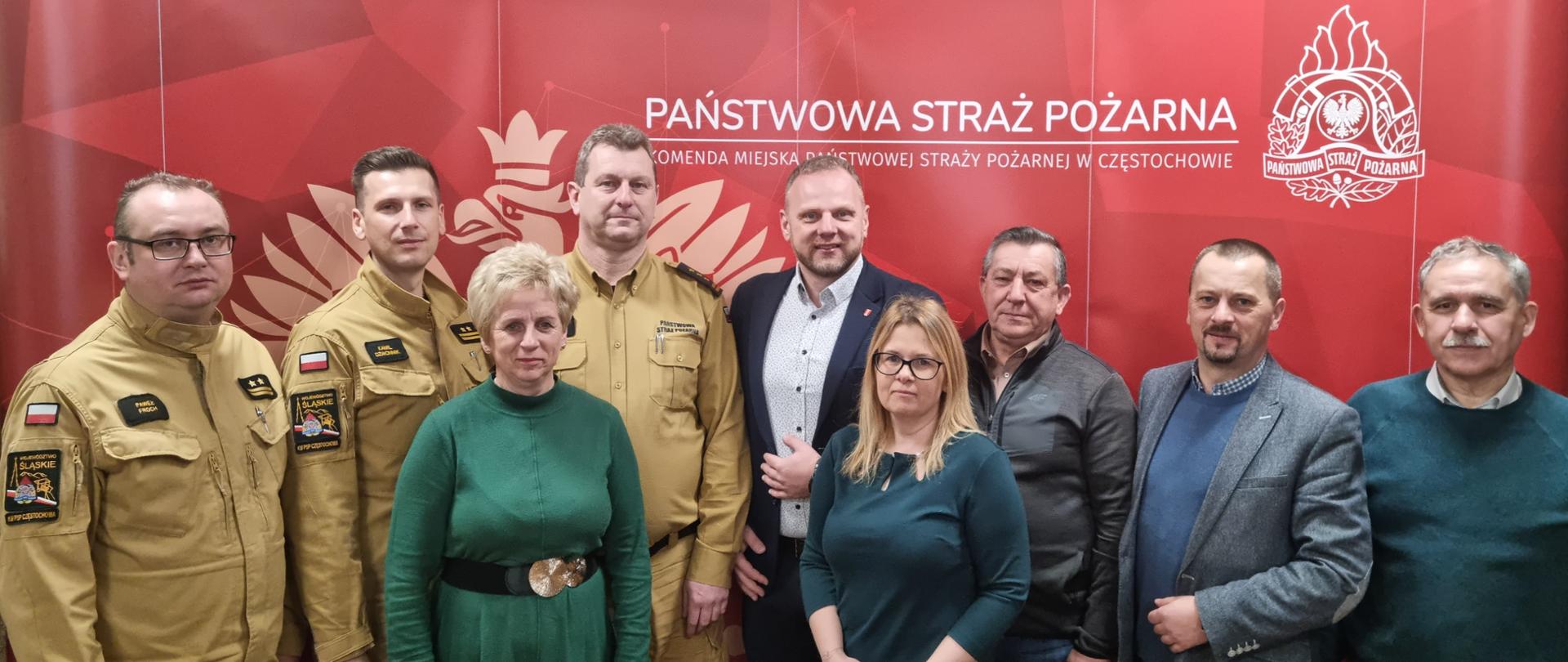 Zdjęcie grupowe uczestników posiedzenia Komisji Samorządności i Bezpieczeństwa Powiatu Częstochowskiego stojący na tle banera reklamowego Komendy Miejskiej Państwowej Straży Pożarnej w Częstochowie.