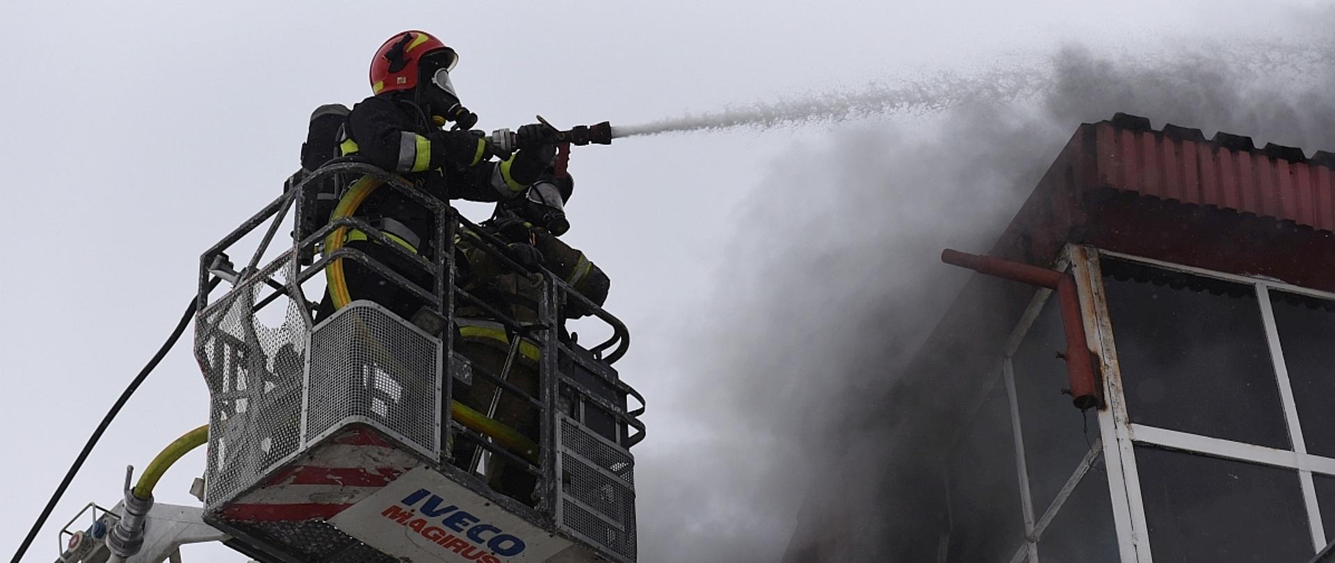Na zdjęciu widać dwóch strażaków w koszu drabiny mechanicznej gaszących pożar hali magazynowej.