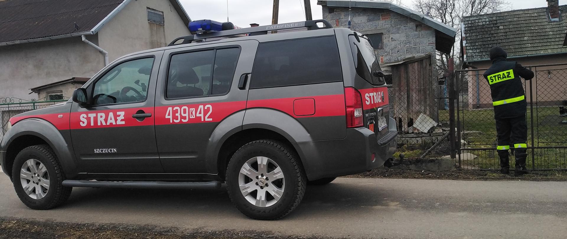 Zdjęcie przedstawia na pierwszym planie samochód operacyjny terenowy Straży Pożarnej, widok od boku, oraz po prawej stronie Strażak wrzucający ulotkę do skrzynki pocztowej w bramie domu jednorodzinnego.