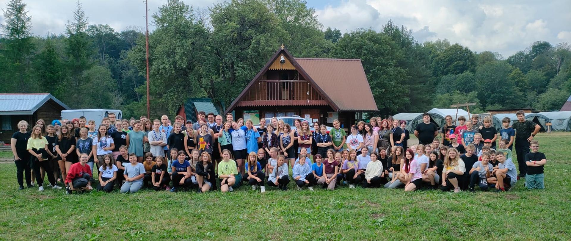 Zdjęcie przedstawia grupę osób (około 100 osób) ustawioną w 2 rzedach. W tle drewniany domek, namioty i drzewa.