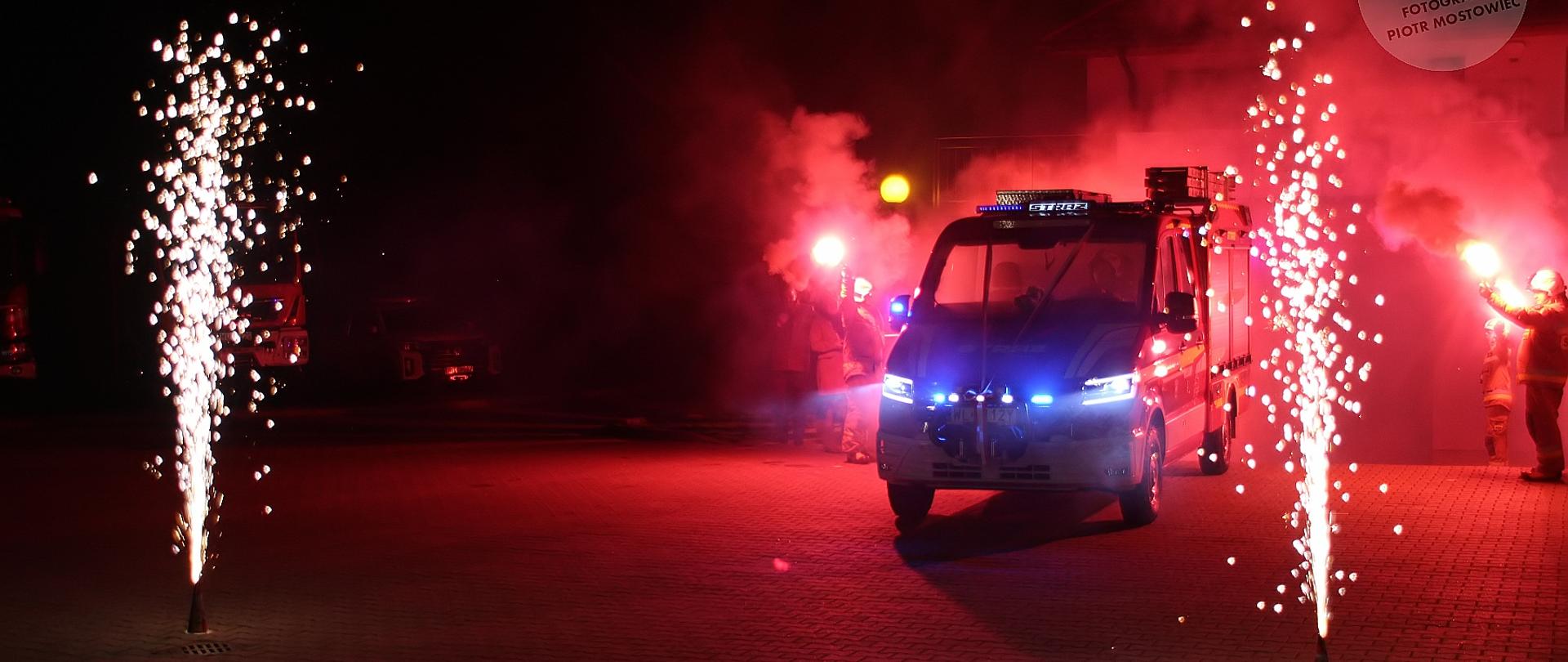 Na zdjęciu widoczna jest nocna scena o bardzo dynamicznym i uroczystym charakterze. Centralnie znajduje się wóz strażacki z włączonymi niebieskimi światłami sygnałowymi, wyjeżdżający lub stojący na placu przed remizą. Pojazd otoczony jest intensywną czerwoną poświatą dymu i światła, co nadaje całości dramatyczny klimat.
Po obu stronach wozu odpalone są fontanny iskier (fajerwerki naziemne), które tworzą świetlistą „bramę”, przez którą przejeżdża samochód. W tle widać strażaków w umundurowaniu ochronnym, trzymających czerwone flary, z których unosi się gęsty dym. Ich sylwetki są częściowo rozświetlone przez ogień i światła pojazdu.
Całość sprawia wrażenie ceremonialnego powitania lub pożegnania – scena jest efektowna, pełna kontrastów między ciemnością nocy a intensywną czerwienią, bielą iskier i niebieskimi światłami uprzywilejowanego pojazdu.