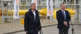 Wicewojewoda opolski oraz prezes Gaz System S.A. na otwarciu obiektu. Mężczyźni stoją przed mikrofonami. Za nimi widać tłocznię - ciąg dużych rur. 