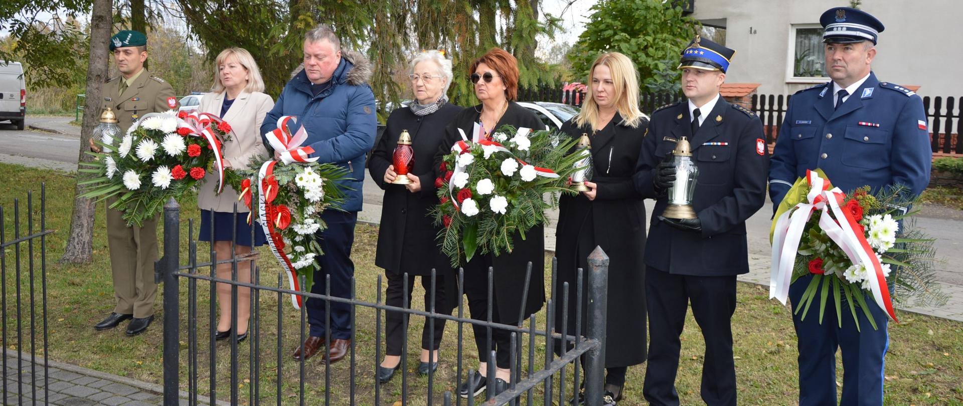 Zdjęcie przedstawia osoby z wiązankami kwiatów oraz zniczami na tle drzew i budynku.