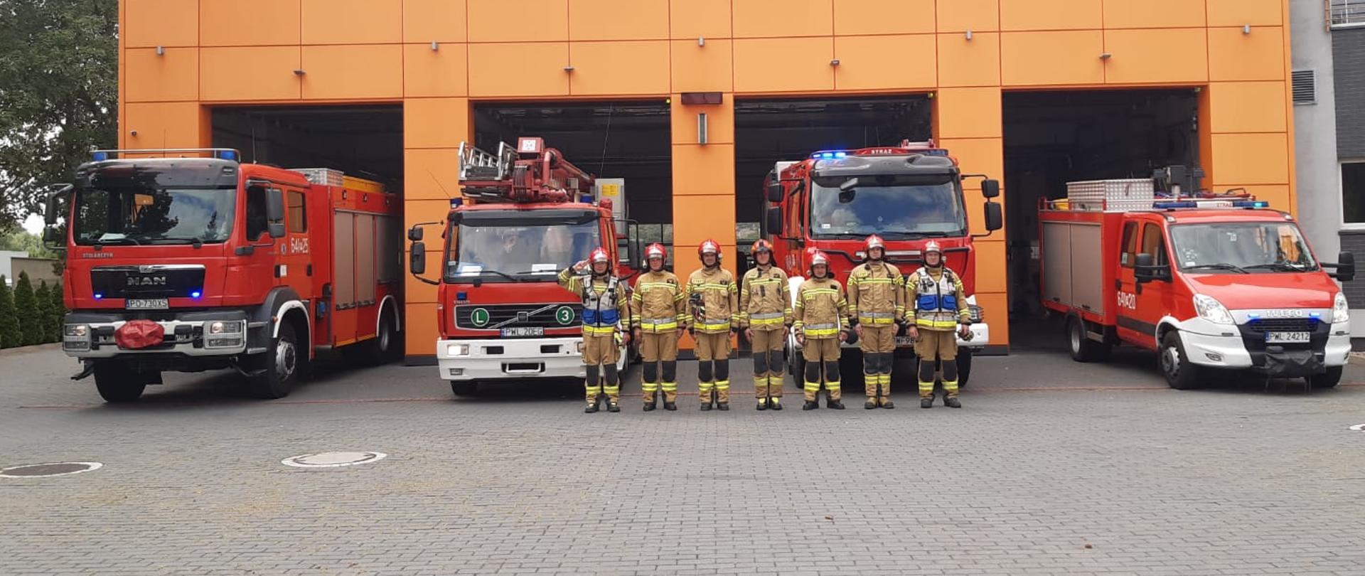 Strażacy stoją przed budynkiem Komendy, ubrani w ubrania specjalne. Oddają hołd powstańcom warszawskim. W tle stoją auta bojowe z włączonymi sygnałami dźwiękowo-świetlnymi.