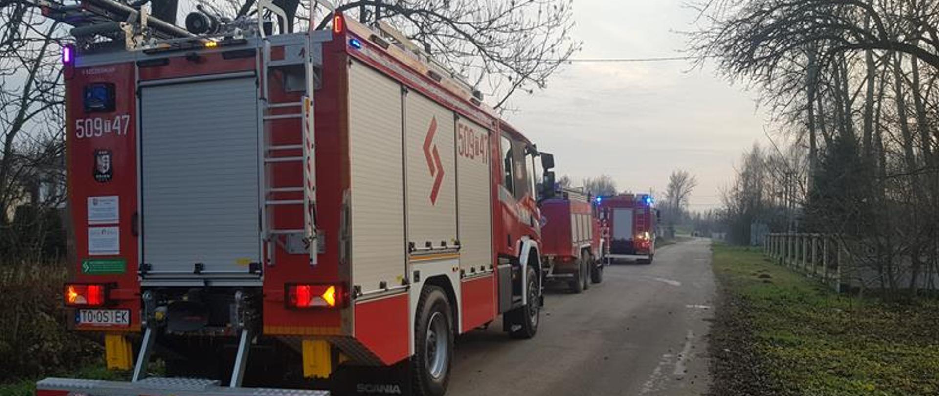 Trzy pojazdy pożarnicze jeden za drugim ustawione są na drodze asfaltowej obok miejsca pożaru. 