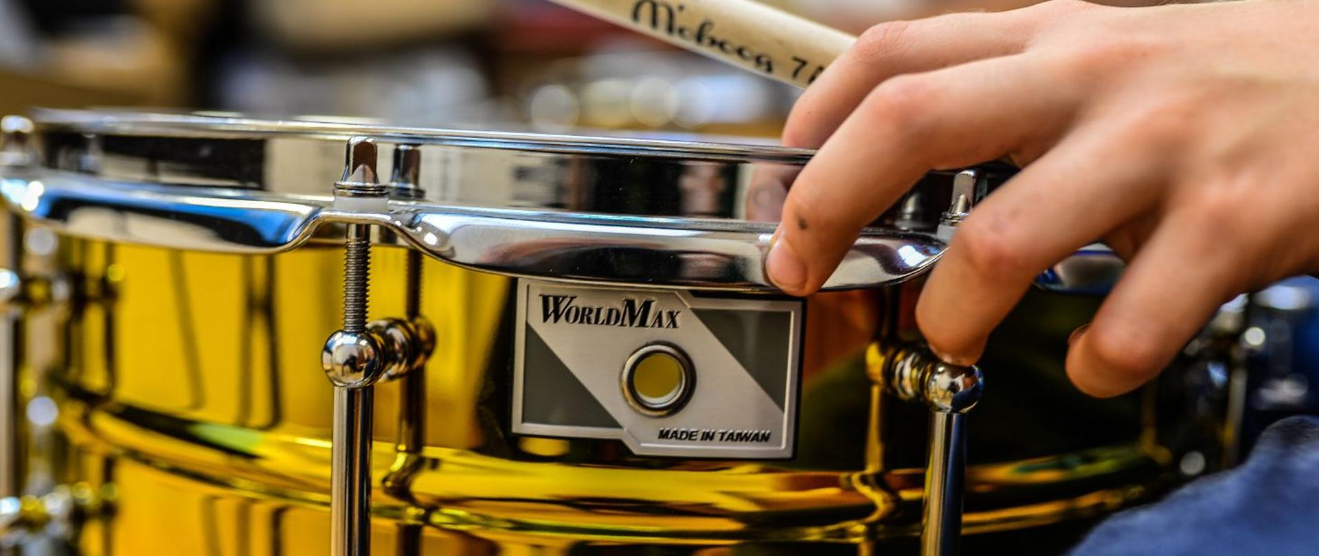 kolorowe zdjęcie przedstawia bok złotego werbla, po lewej stronie dłonie trzymające pałki do perkusji