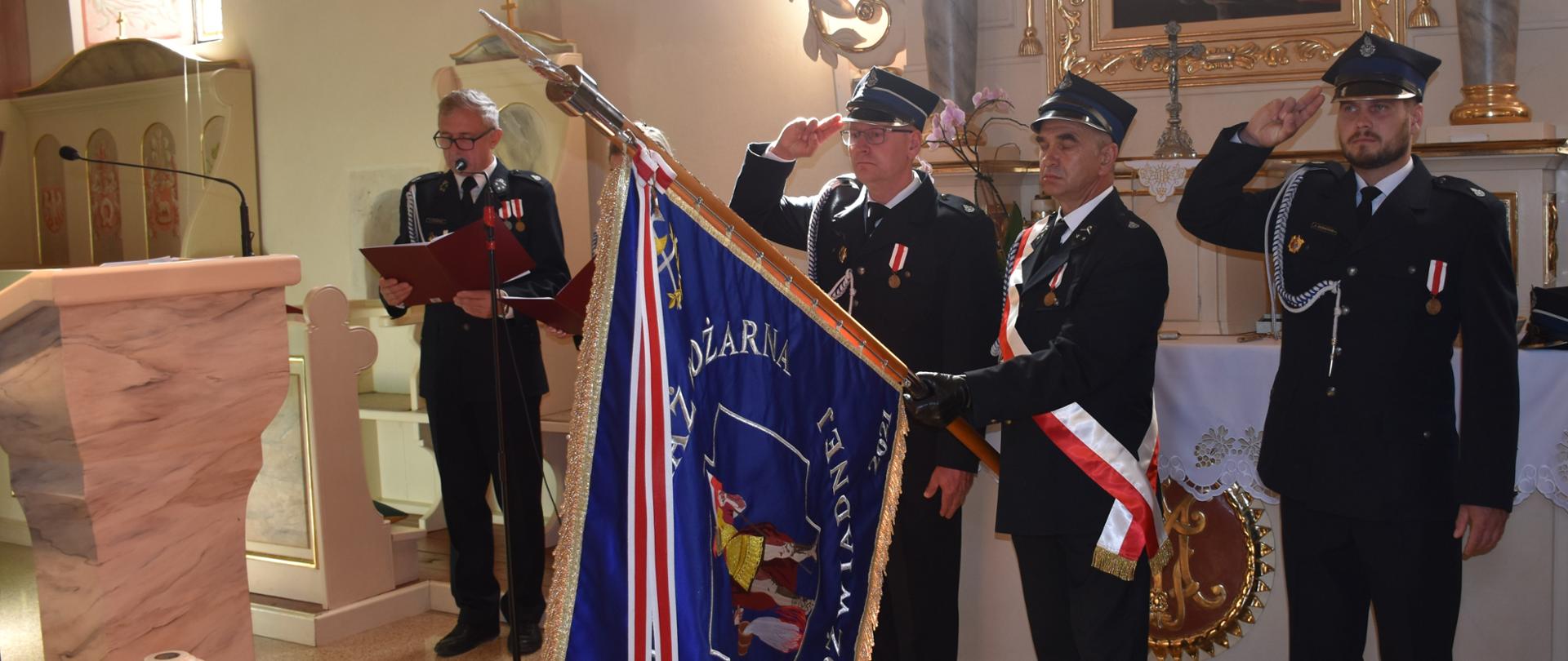 Poczet sztandarowy OSP salutuje sztandarem w kościele. Z lewej strażak OSP czyta tekst.