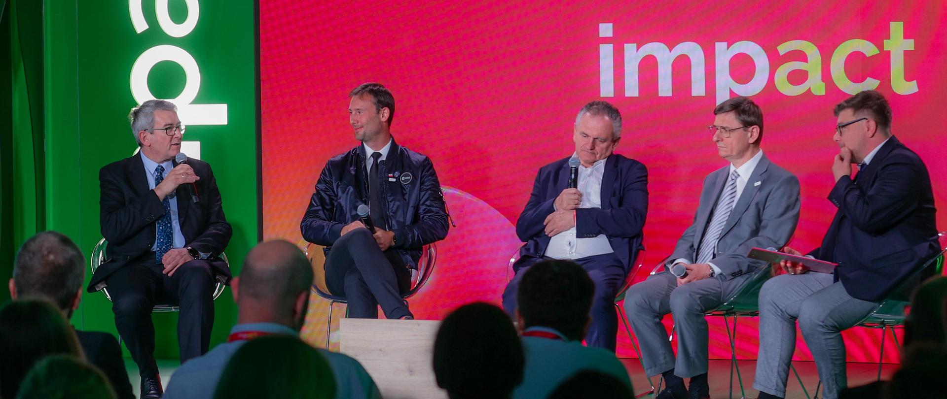 Pod czerwono-zieloną ścianą z napisem Impact'23 na fotelach siedzi pięciu mężczyzn, na skraju z lewej siedzi wiceminister Murdzek który mówi do trzymanego w ręku mikrofonu.