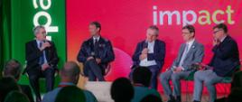 Pod czerwono-zieloną ścianą z napisem Impact'23 na fotelach siedzi pięciu mężczyzn, na skraju z lewej siedzi wiceminister Murdzek który mówi do trzymanego w ręku mikrofonu.