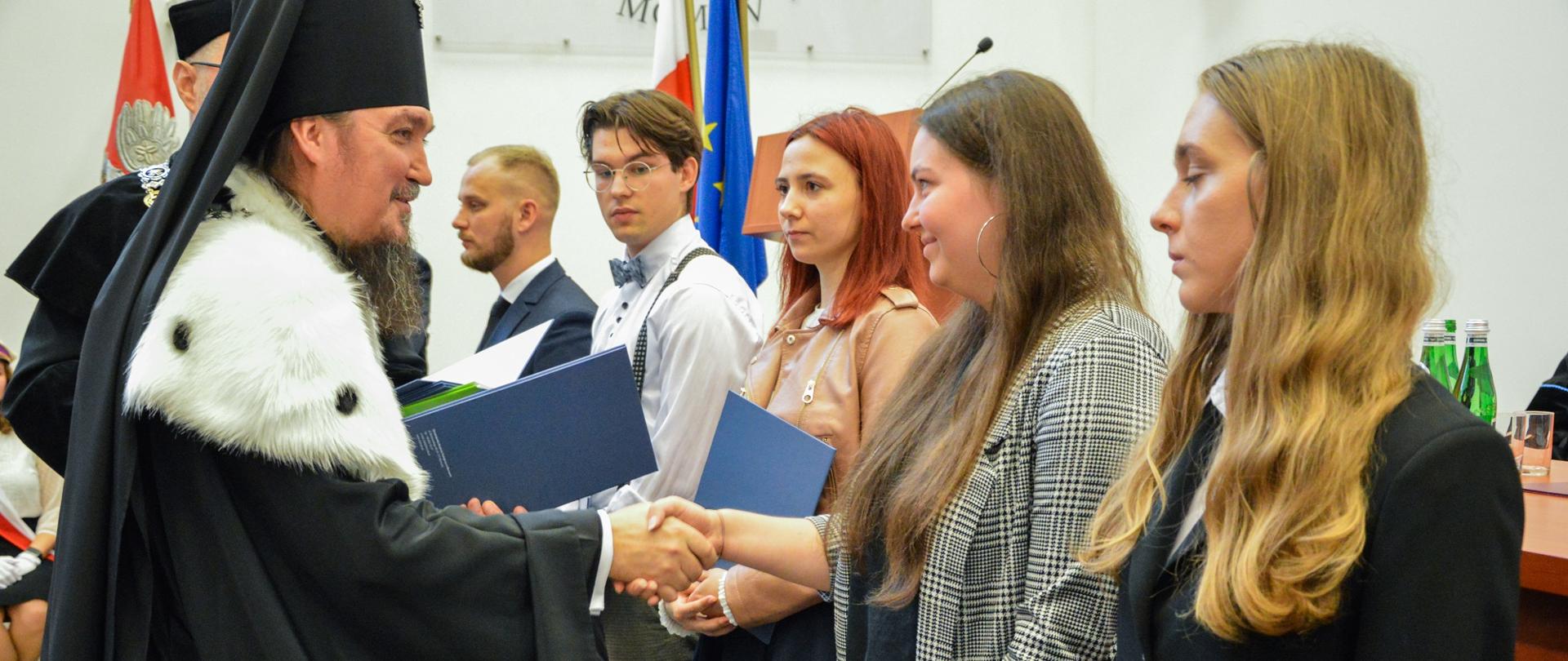 Mężczyzna w czarnym ceremonialnym stroju ściska rękę młodej dziewczynie, stojącej w grupie innych dziewczyn.