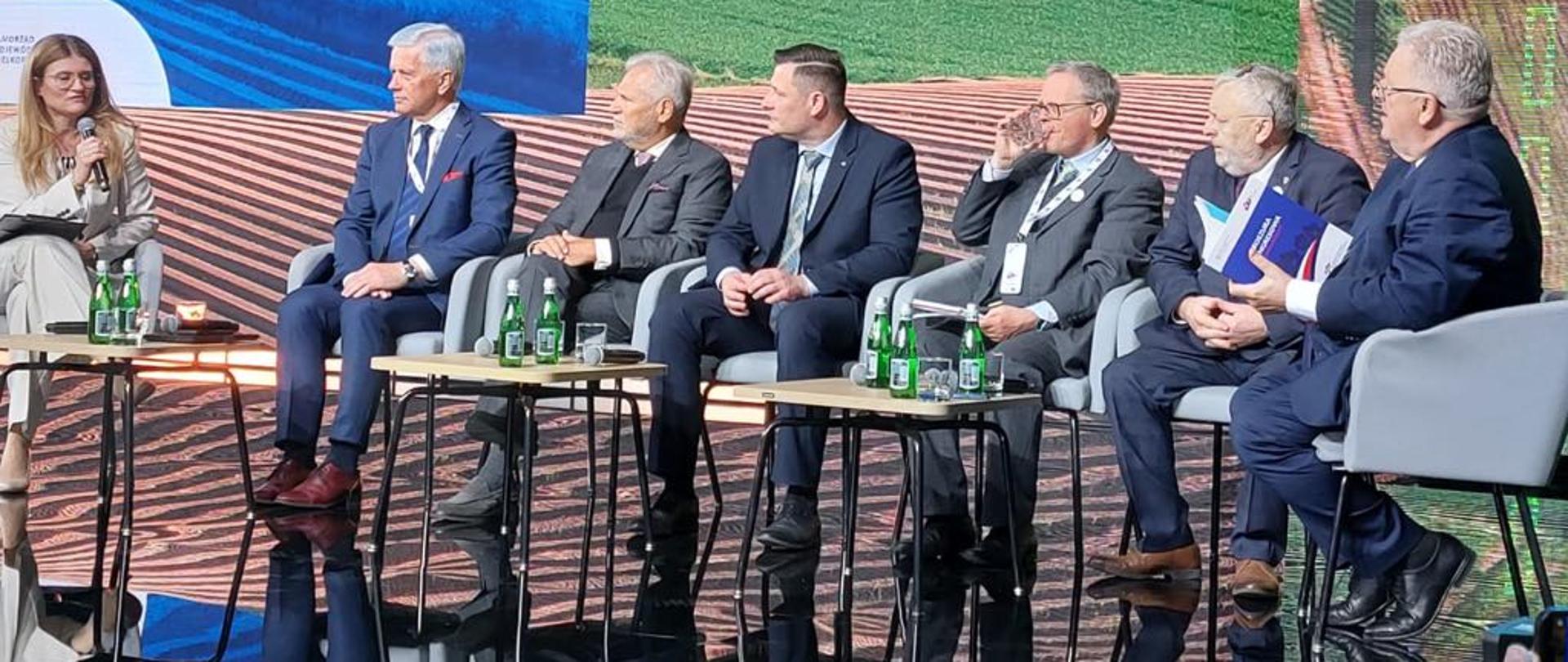 Fotografia przedstawia kobietę z mikrofonem po lewej stronie w towarzystwie 6 mężczyzn ubranych w garnitury. W tle wizerunek pola uprawnego.