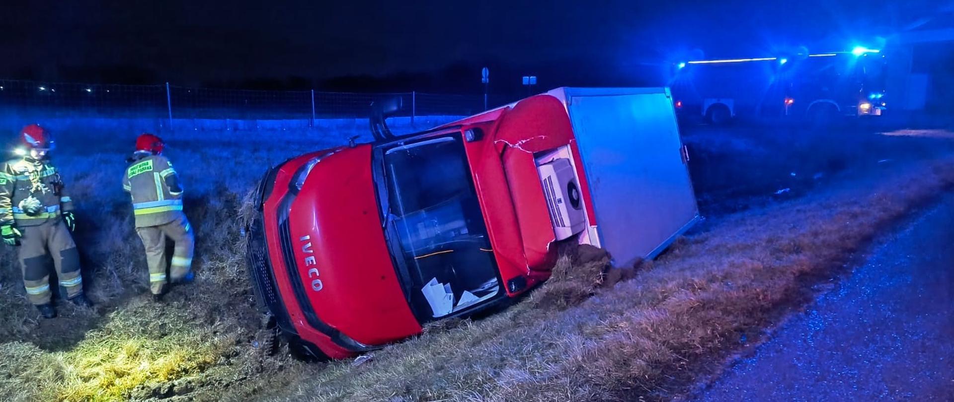 Samochód dostawczy Iveco leży na poboczu na lewym boku, obok dwóch strażaków a w tle samochód GBA 3/16