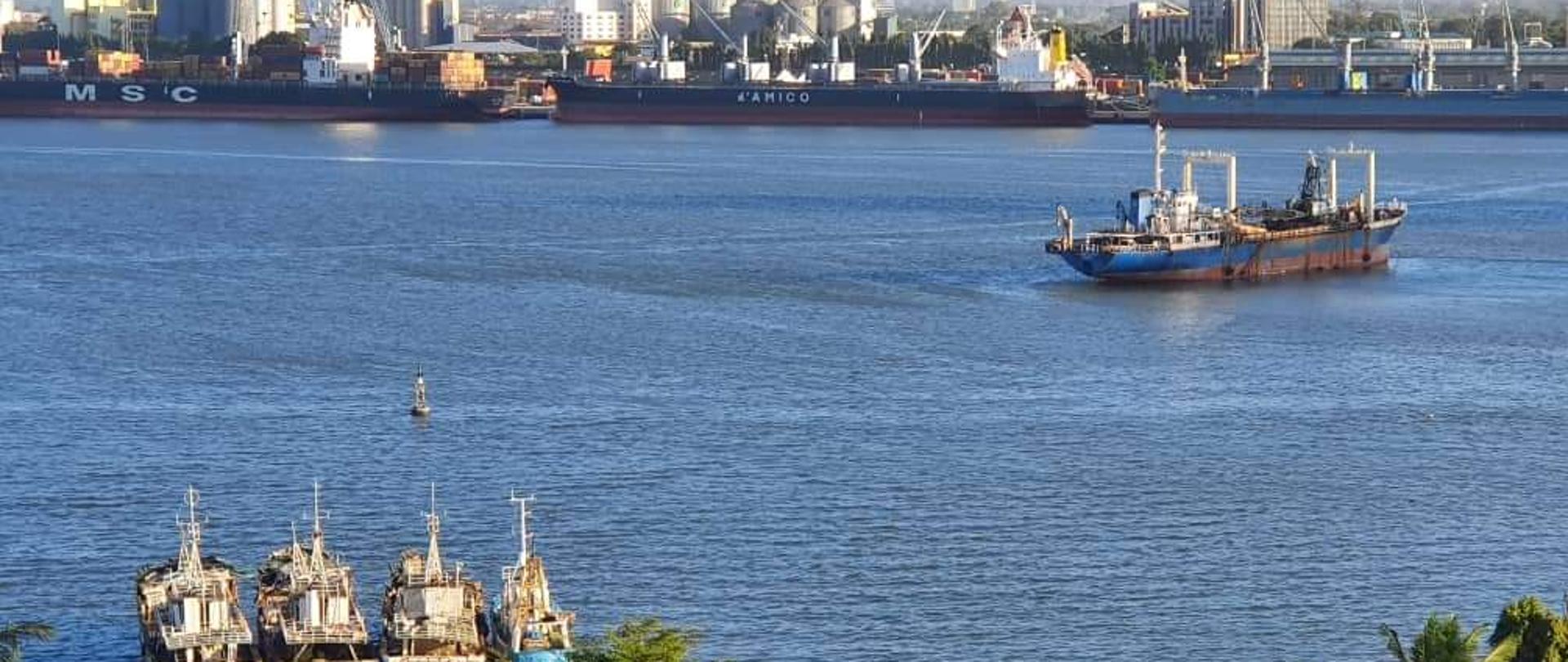 na wodzie 5 statków oraz kilka statków cargo, w tle zabudowania przemysłowe i miejskie