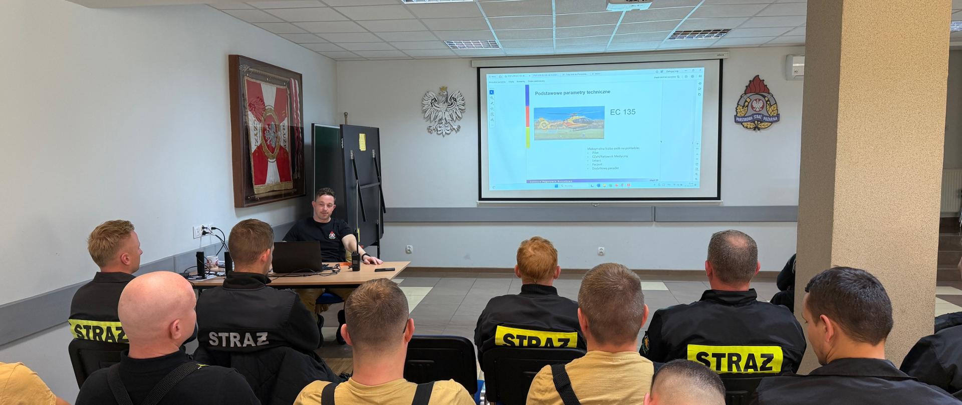 Druhowie Ochotniczych Straży Pożarnych z powiatu mławskiego uczestniczą w szkoleniu z zakresu współdziałania z Lotniczym Pogotowiem Ratunkowym. Instruktor PSP omawia podstawowe parametry śmigłowca LPR EC 135, korzystając z prezentacji multimedialnej wyświetlanej na ekranie.