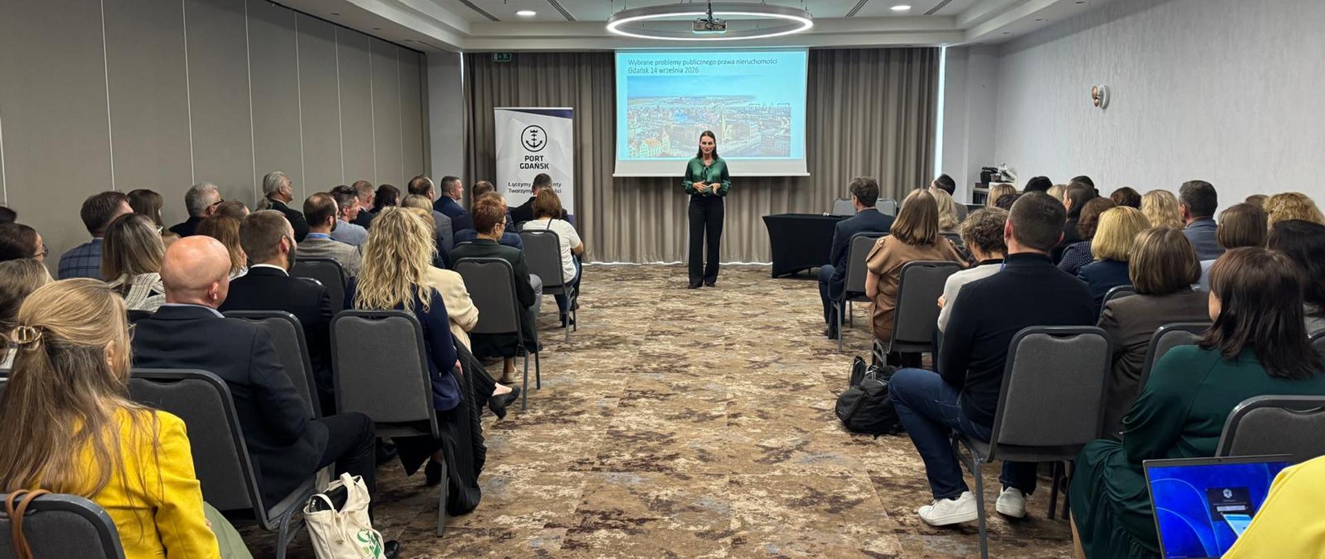 Prelegentka w zielonej bluzce i czarnych spodniach stoi przed ekranem projekcyjnym w sali konferencyjnej, przemawiając do licznie zgromadzonej publiczności siedzącej na krzesłach.