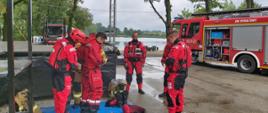 Na zdjęciu strażacy przygotowujący się do działań na wodzie. W tle zbiornik wodny oraz samochód strażacki.
