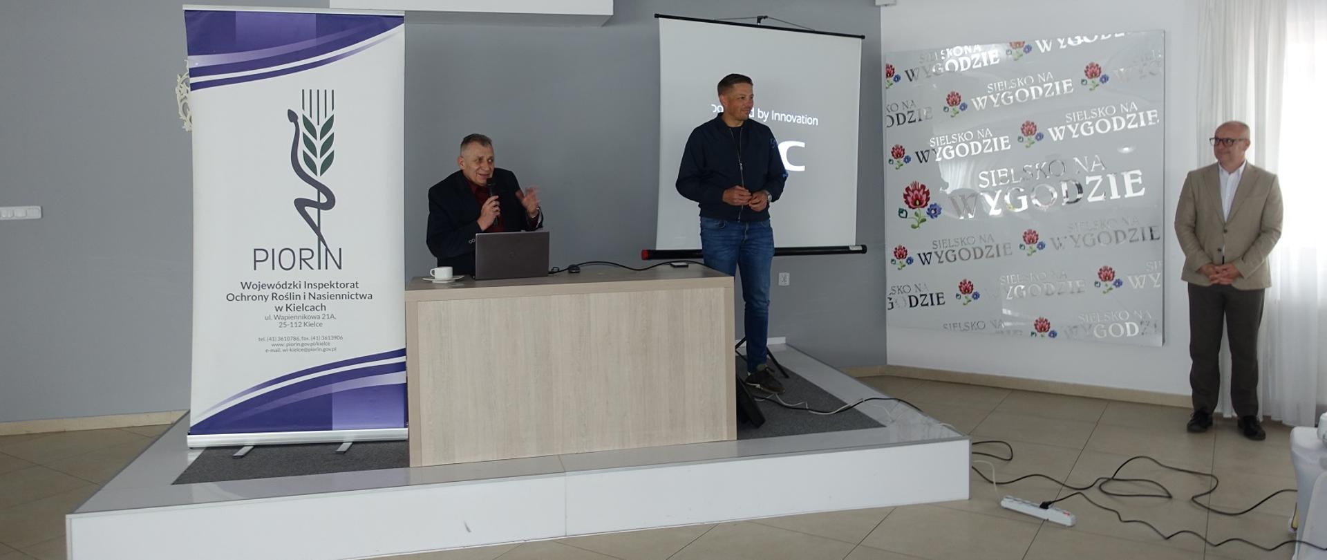 Podium sali wykładowej. Świętokrzyski Wojewódzki Inspektor Ochrony Roślin i Nasiennictwa wygłasza przemówienie do pracowników. Zastępca Pan Marcin Chłodnicki oczekuje na przejęcie mikrofonu. Po lewo widoczny jest roll-up z logo PIORiN. Pan wykładowca stojący po prawej stronie przysłuchuje się temu co mówi Pan Edmund Kaczmarek.