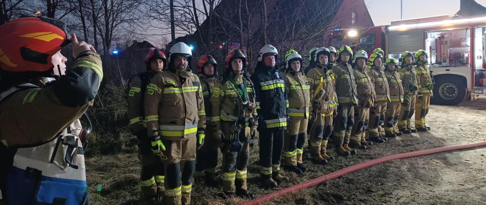 Strażacy oddają hołd zmarłemu koledze, stojąc w rzędzie na baczność.