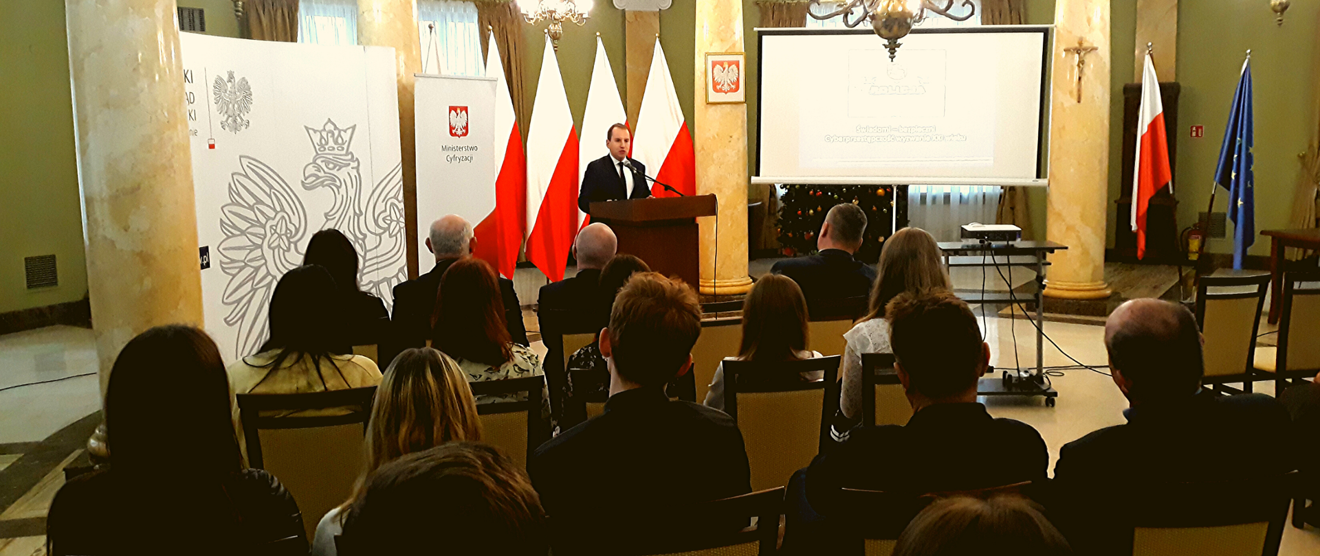 Adam Andruszkiewicz przy mównicy. W tle ścianka Ministerstwa Cyfryzacji i polskie flagi. Ujęcie zza pleców pozostałych uczestników spotkania, którzy siedzą naprzeciw ministra.