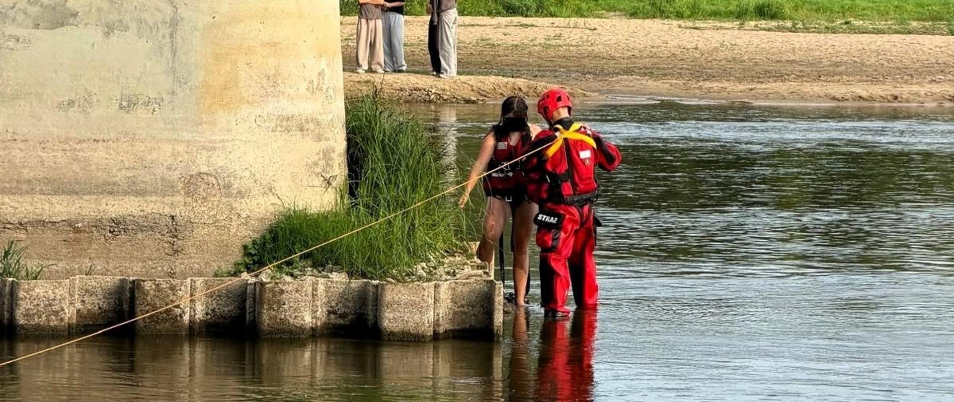 Betonowy filar mostu drogowego stoi w rzece. Na podstawie filaru stoi strażak ubrany w suchy skafander i dziewczynka z założona kamizelką asekuracyjną. W tle piaszczysta plaża, rosnąca dalej trawa oraz osoby znajdujące się na plaży i trawie.