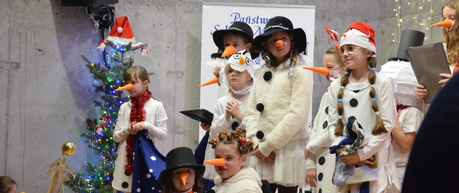 Grupa dzieci przebranych za bałwanki stoi na scenie, choinka, świąteczne dekoracje