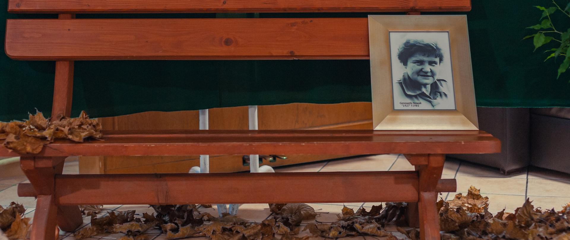 Zdjęcie fragment dekoracji. Drewniana ławka na tle ciemnej tkaniny. Po prawej wsparta o oparcie fotografia Genowefy Nowak w ramce. Wokół rozrzucone jesienne liście.