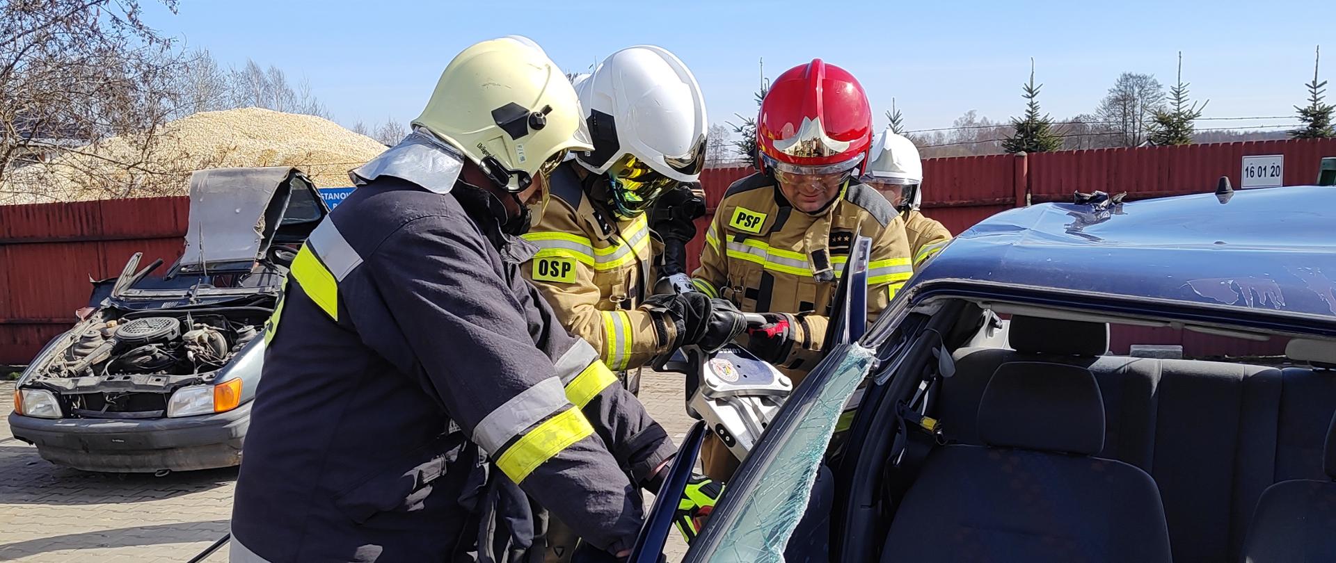 Dowódca zmiany z PSP wyjaśnia druhom OSP otwarcie drzwi przy użyciu rozpieracza ramieniowego. 