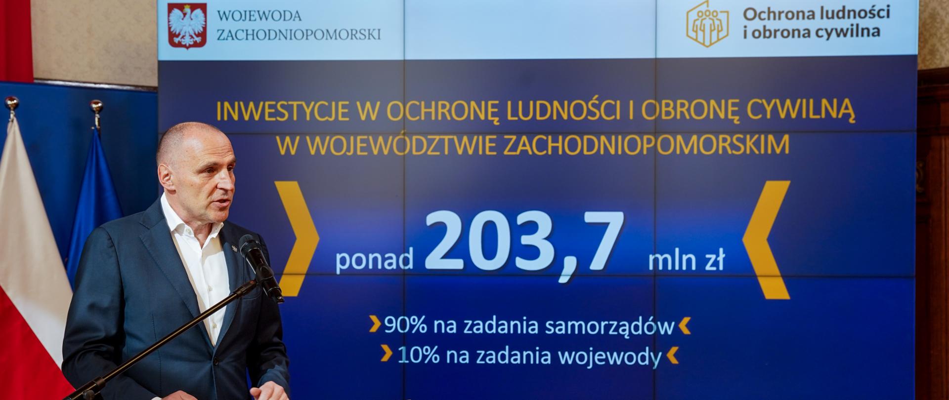 wojewoda na tle planszy z kwotą 203,7 miliona złotych na inwestycje w ochronę ludności i obronę cywilną