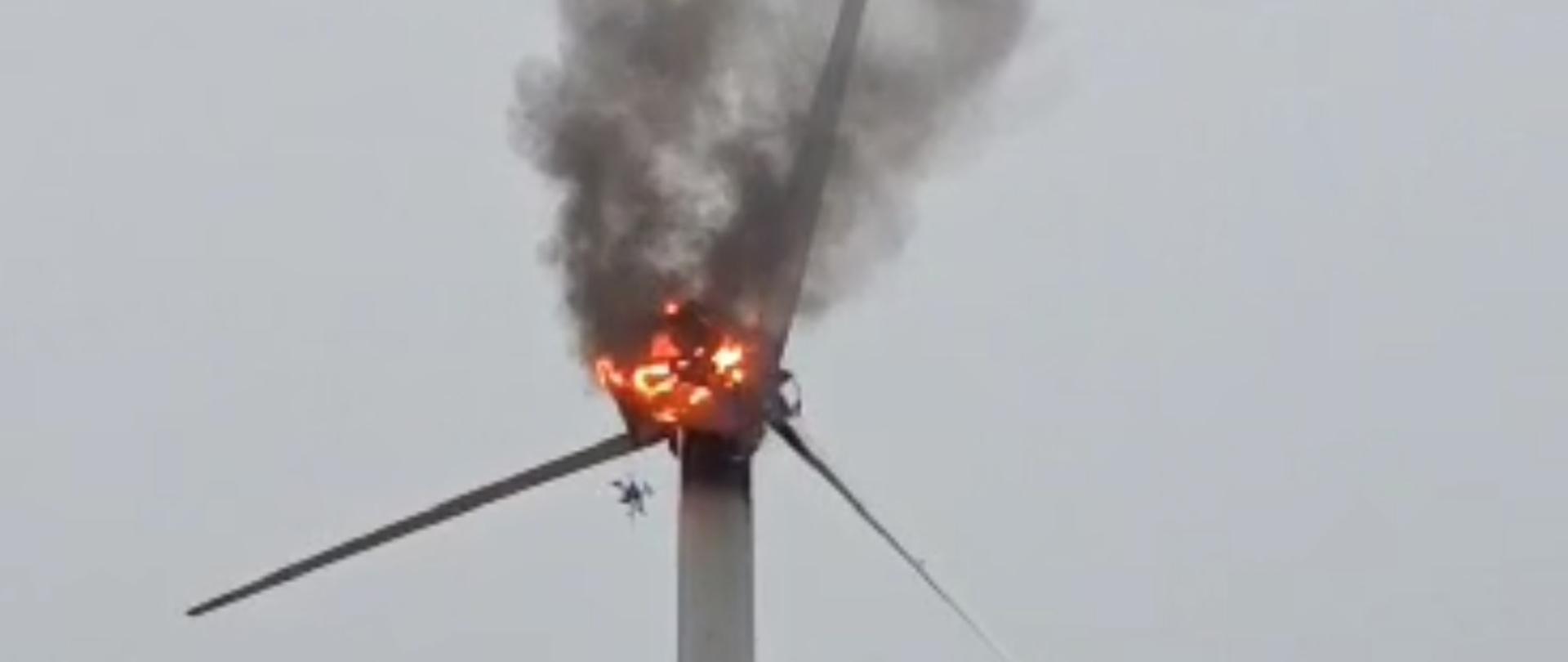 Łąka, na drugim planie wieża na której zainstalowana jest gondola wiatraka ze śmigłami z której wydobywa się dym i ogień